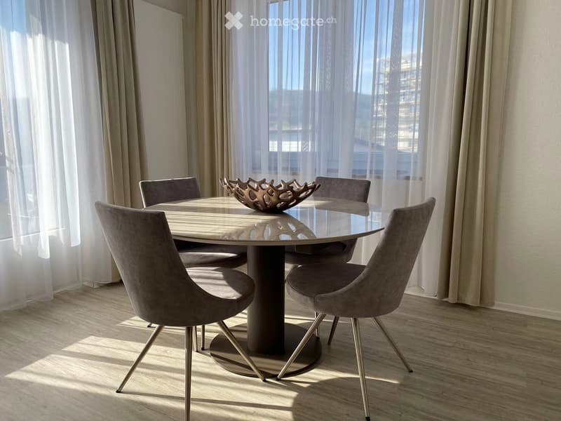 Dining room with a round glass table, four upholstered chairs, and a decorative bowl on the table. Large windows with sheer curtains allow natural light to fill the space.