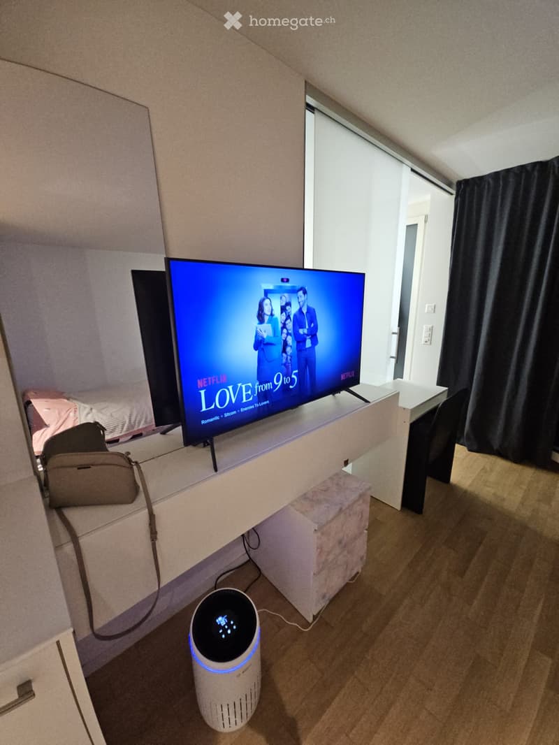 A modern living room with a large flat-screen TV displaying a Netflix show, a sleek white table, a chair, a handbag, and an air purifier on the floor.