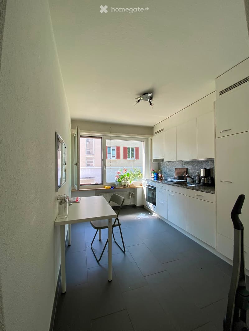 Modern kitchen with white cabinets, dark tiled floor, and a small dining area. Features include an oven, fridge, coffee machine, sink, and a small table with chairs.