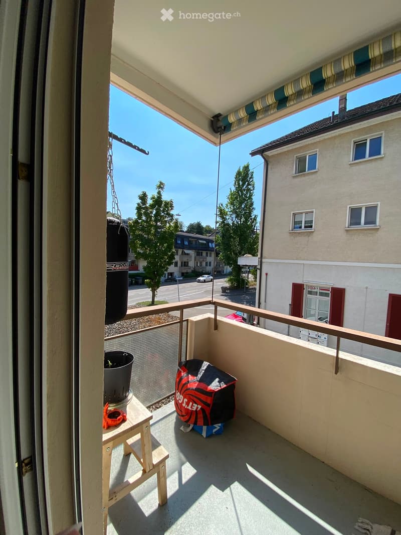 Balcony with a small wooden stool, a potted plant, and a backpack, overlooking a street with buildings and trees.