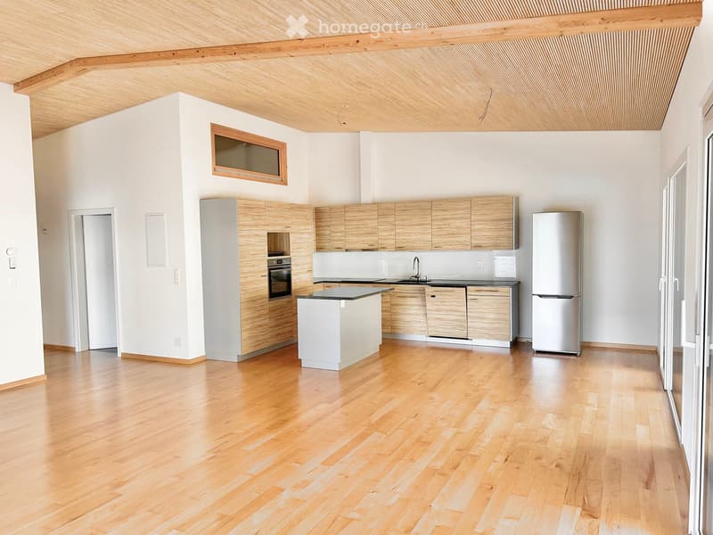 Modern kitchen with light wooden cabinets, stainless steel appliances, and a central island. The space features a polished wooden floor and large windows.