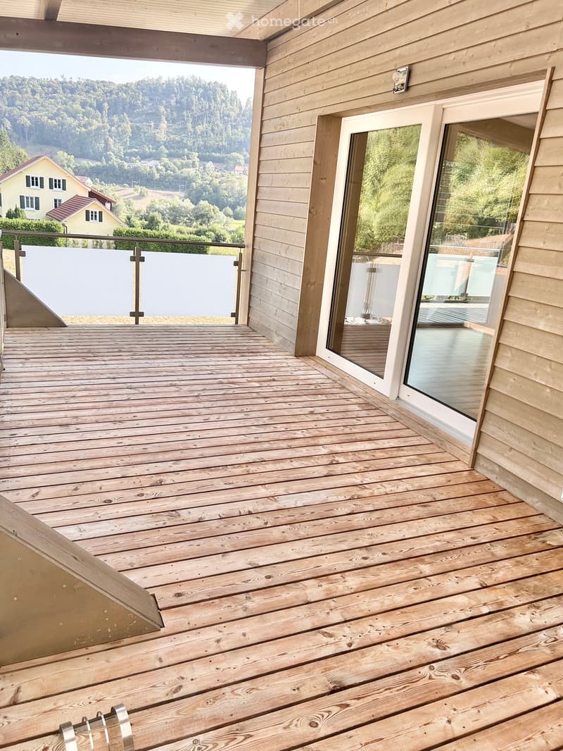 Wooden balcony with glass doors, metal railing, and scenic view of greenery and houses.