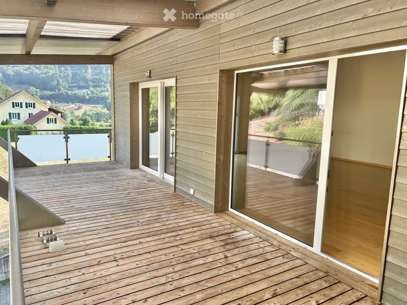 Wooden deck with glass sliding doors, metal railing, and scenic view of greenery and houses.
