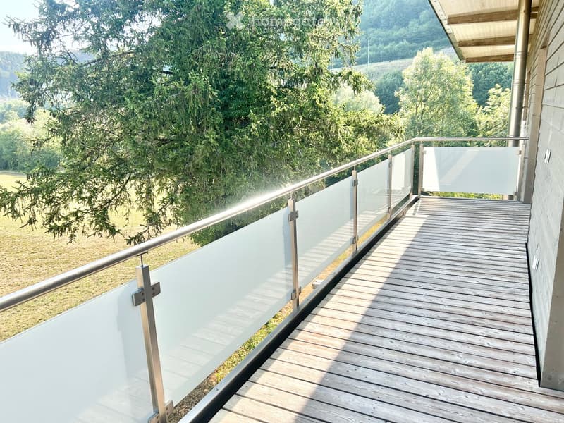 Wooden deck with glass railing, overlooking a green landscape with trees and hills.