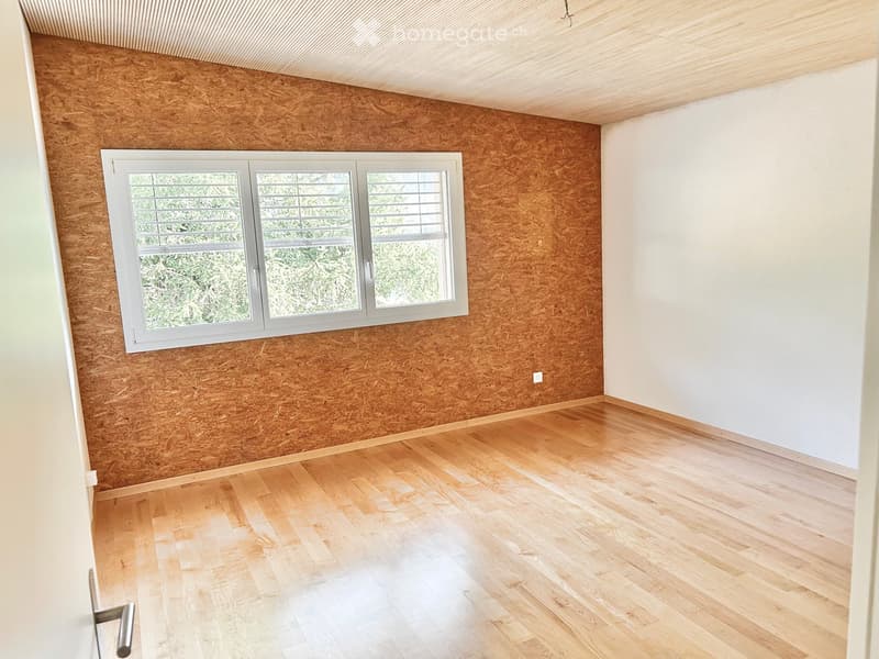 Empty room with wooden flooring, white walls, and a large window with blinds. The walls feature a textured wood panel design on one side.