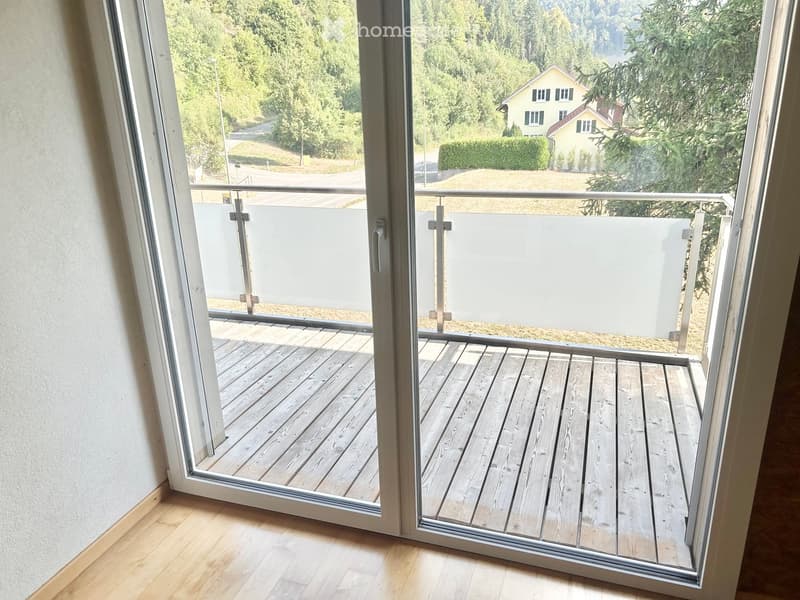 Balcony with wooden decking, glass sliding doors, and a view of a green landscape with trees and a house.