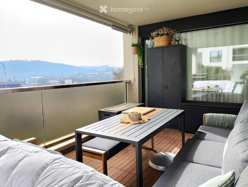 Balcony with wooden decking, grey sofa, small table, stools, cabinet, and potted plants, offering a scenic view of the city and hills.