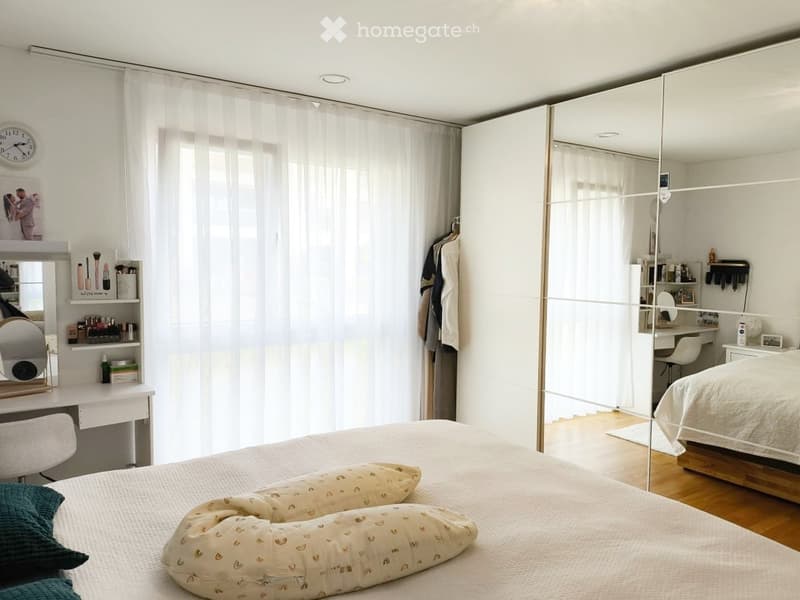 Modern bedroom with a large bed, white wardrobe, desk with chair, and a shelf with various items. The room features a large window with sheer curtains and a wooden floor.