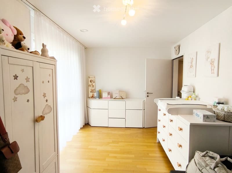 Bright, modern children's bedroom with a large wardrobe, changing table, storage unit, and decorative toys on top of the wardrobe. The room features light wooden flooring and white walls.