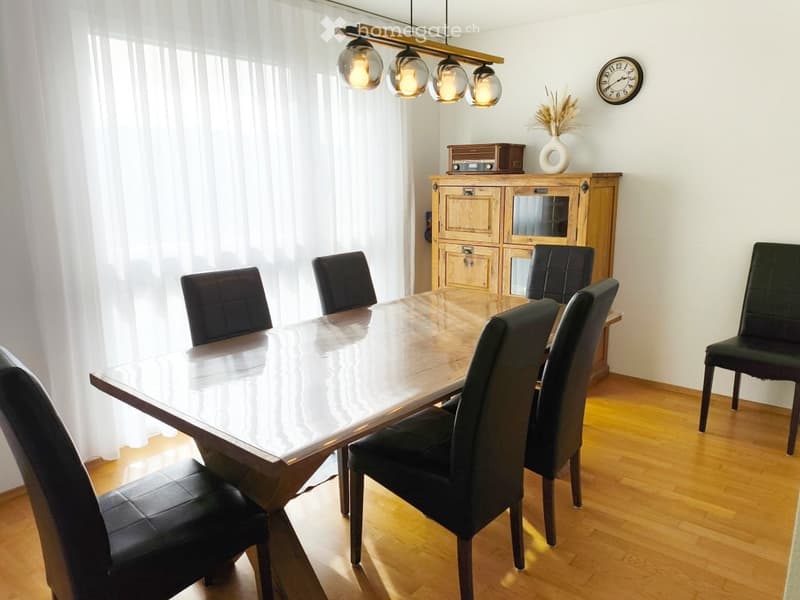 Dining room with a wooden dining table, six black leather chairs, a wooden cabinet with glass doors, a clock on the wall, and a vase with dried flowers on the cabinet.