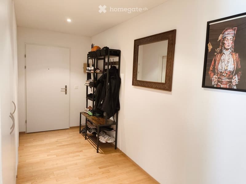 A clean, modern hallway with light wooden flooring, white walls, a coat rack with shoes and jackets, a large mirror, and two framed artworks.