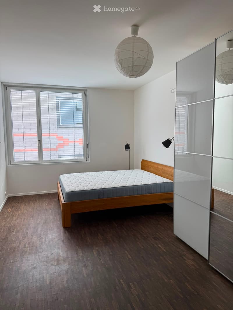 Modern bedroom with wooden flooring, a single bed with a grey mattress, a large mirrored wardrobe, and a large window with blinds.