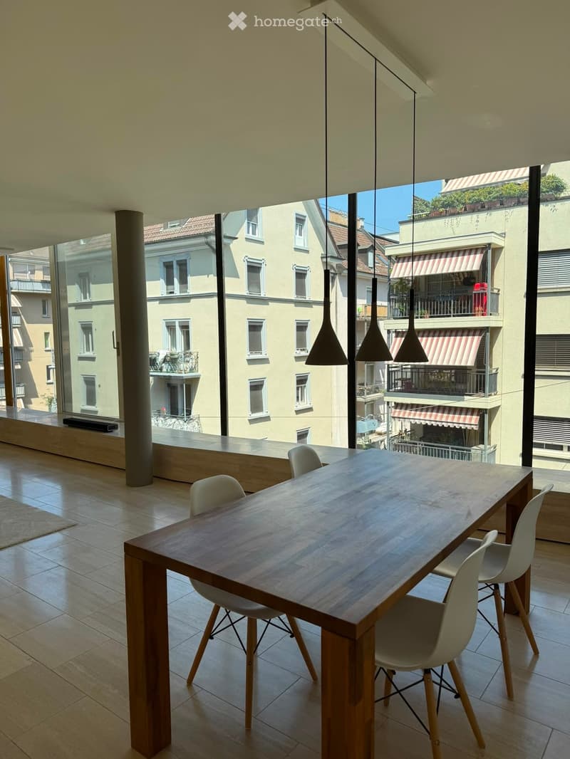 Bright, modern dining area with a long wooden table and white chairs, large windows overlooking a cityscape, and hanging pendant lights.
