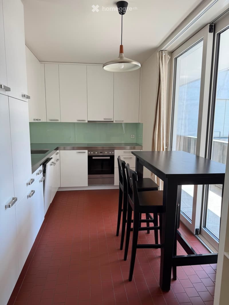 Modern kitchen with white cabinets, stainless steel appliances, red tiled floor, and a black dining table with chairs.