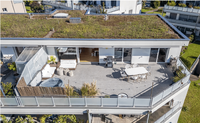 Modern rooftop terrace with a hot tub, seating area, dining table, and barbecue, surrounded by greenery and glass railings.