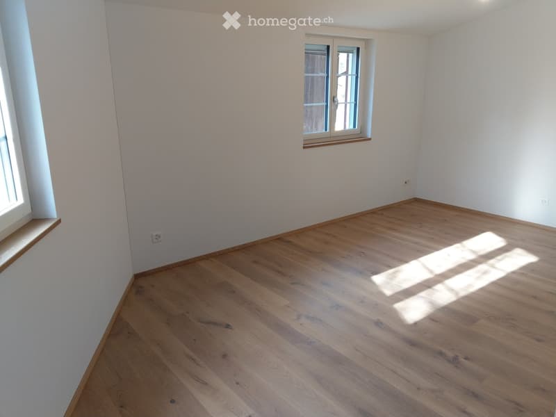 Empty room with light wooden flooring and white walls, featuring two windows with wooden frames and sills.