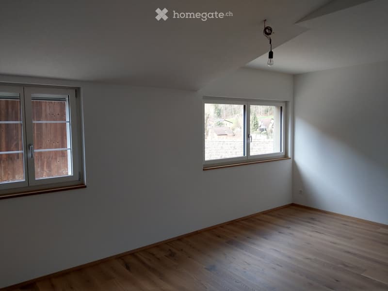 Empty room with wooden flooring, white walls, and two windows, one with a view of a house and trees outside.