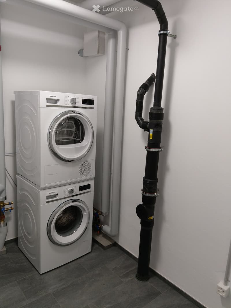 A modern bathroom with a stacked washing machine and dryer unit, white walls, and grey tiled floor.