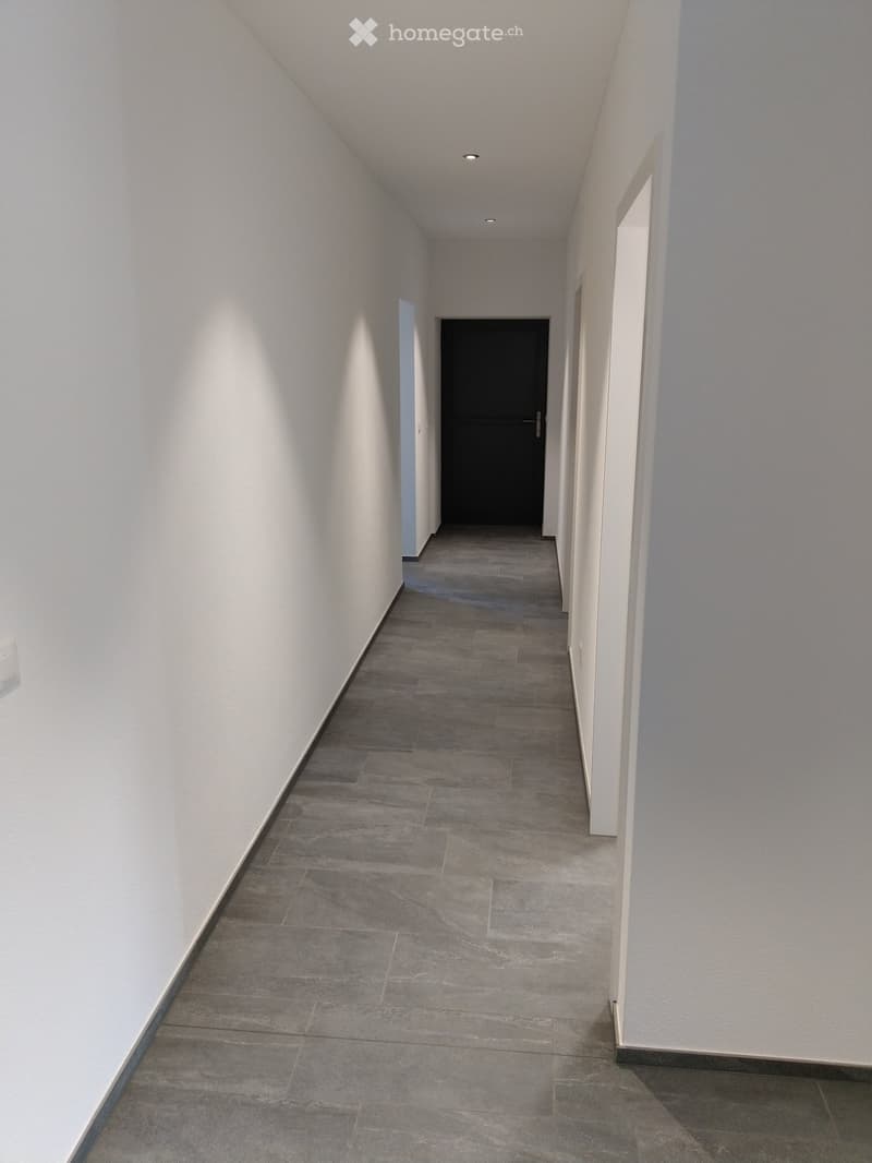 A long, narrow hallway with grey tiled flooring and white walls, featuring a closed black door at the end and recessed ceiling lighting.