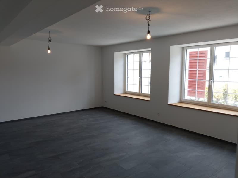 Empty room with grey walls, dark grey tiled floor, two windows with wooden sills, and two hanging light fixtures.