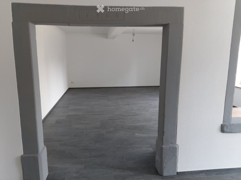 Empty hallway with dark grey tiled floor, white walls, and a concrete archway leading to another room.