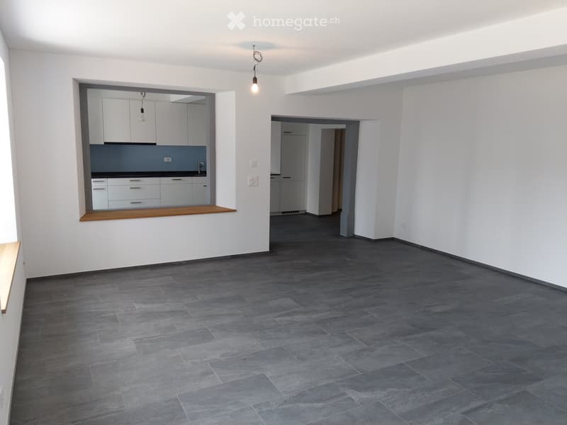 Empty room with grey tiled floor, white walls, and visible kitchen and bathroom areas in the background.