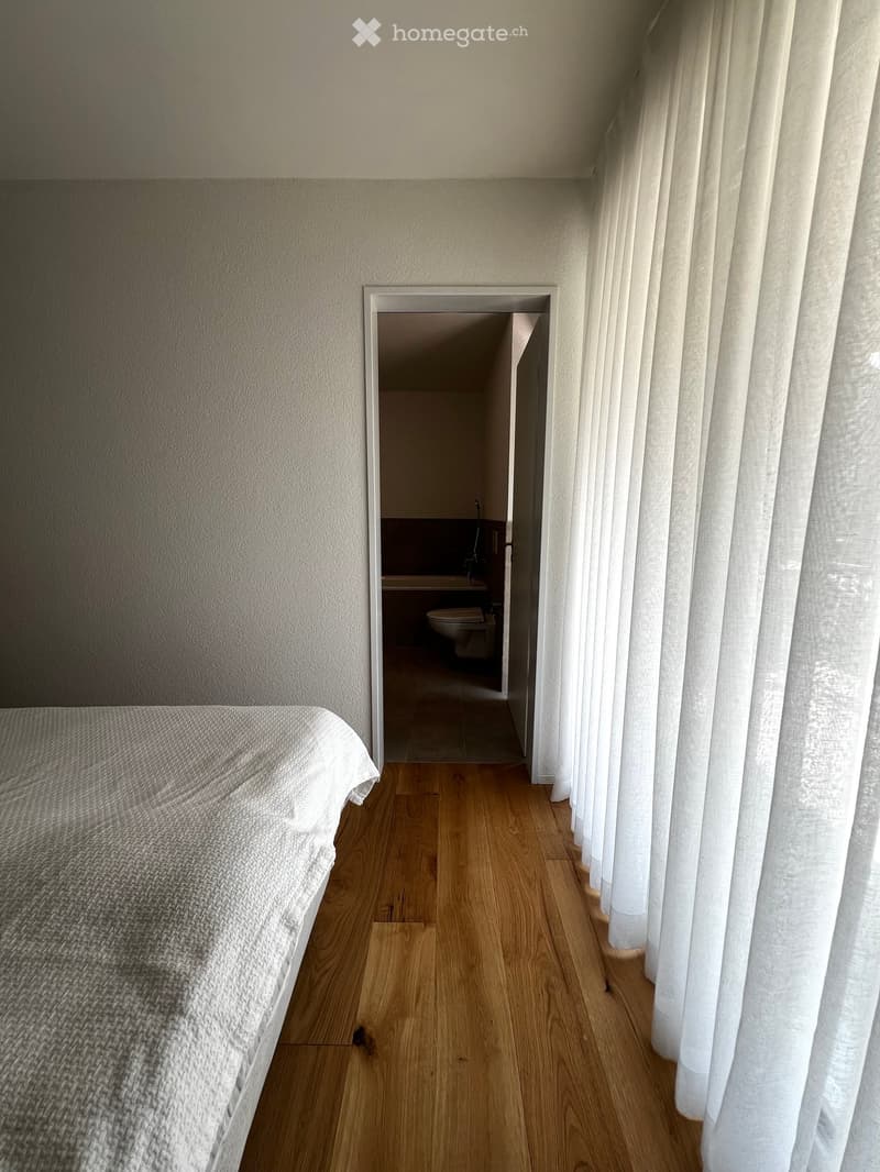 Bedroom with wooden flooring, a neatly made bed on the left, and an open doorway leading to a small bathroom with a toilet and sink.