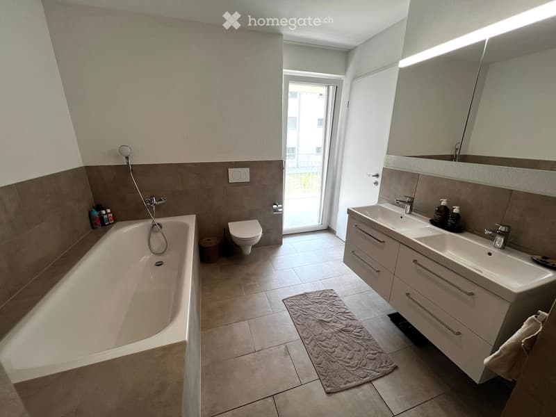 Modern bathroom with a bathtub, toilet, double sink, large mirror, and a small window leading to a balcony.