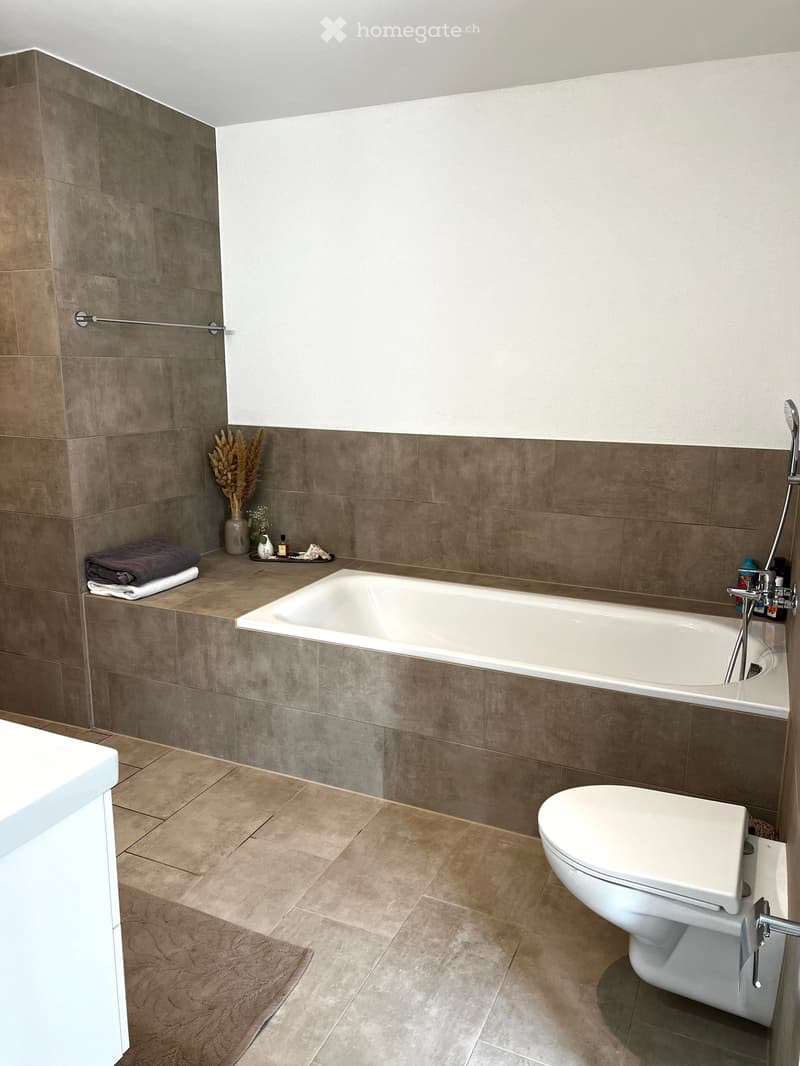 Modern bathroom with a bathtub, toilet, and a towel on a shelf. The walls are tiled in a neutral color scheme, and there is a vase with dried flowers on the shelf.