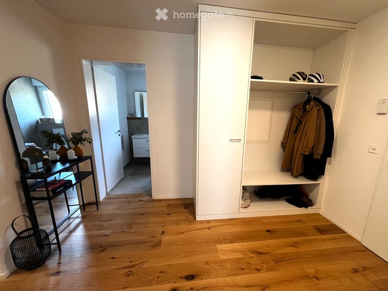A well-lit hallway with wooden flooring, a large built-in wardrobe, a small table with a lamp and vases, and an open doorway leading to a bathroom.