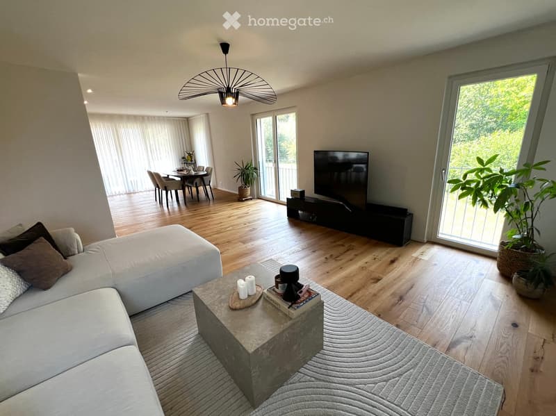 Modern living room with a white sectional sofa, coffee table, TV, dining area, and large windows with sheer curtains.