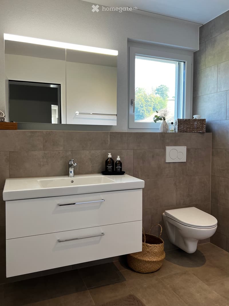 Modern bathroom with a double sink, large mirror, toilet, and wicker basket on the floor. The sink has a sleek faucet and is paired with a modern cabinet. A window with a view of greenery is visible in the background.