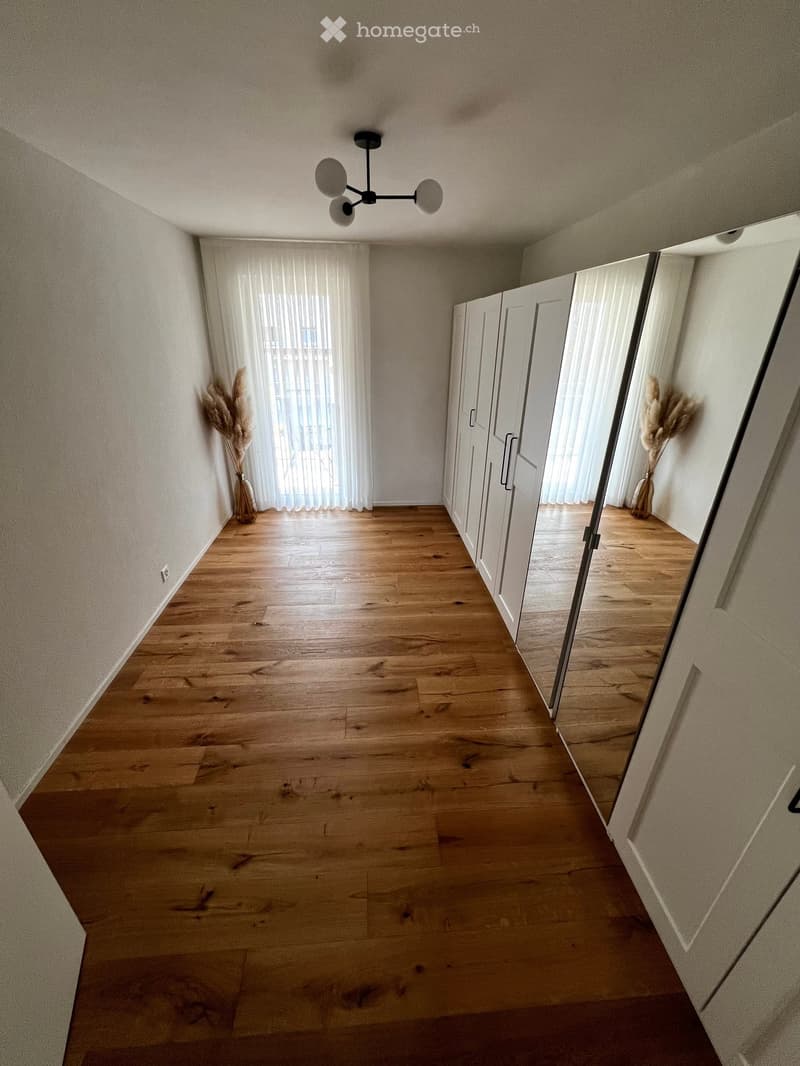 Empty hallway with wooden flooring, white walls, and modern lighting fixture. Two large mirrored wardrobes on one side and a window with sheer curtains on the other.