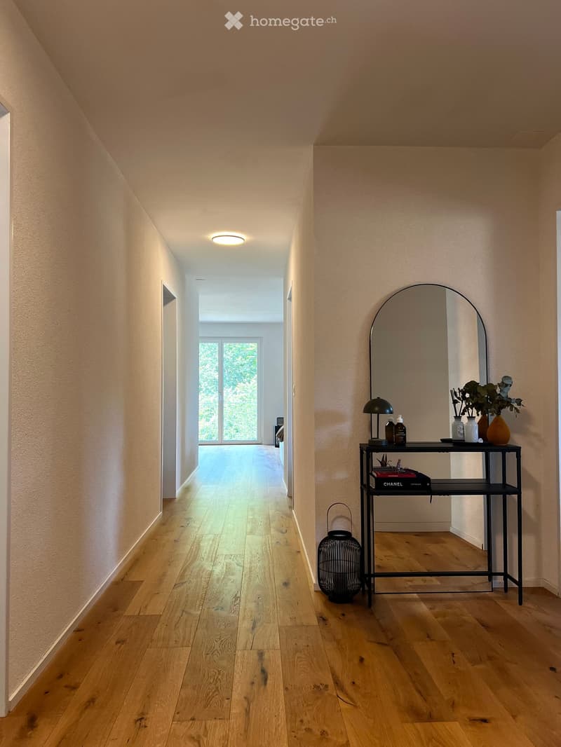 A modern hallway with light wooden flooring, beige walls, and a sleek black console table holding decorative items, a mirror, and a lamp.