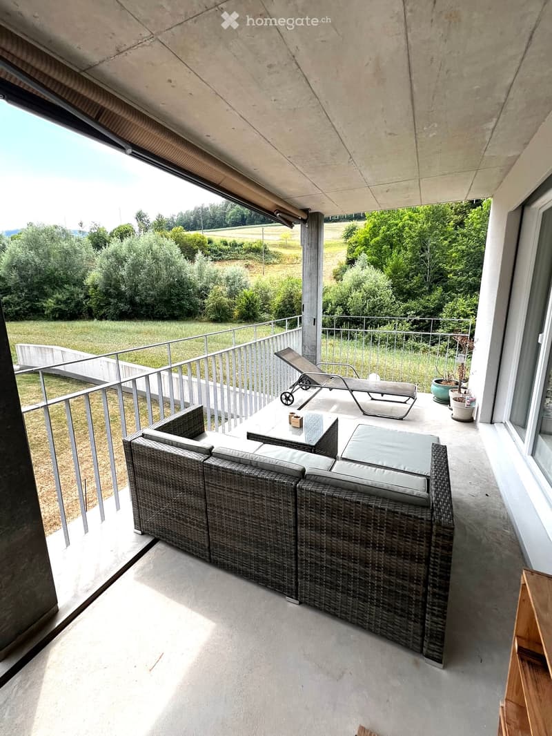 Balcony with a modern design, featuring a wicker sofa, a glass table, a lounge chair, and potted plants, overlooking a green landscape.