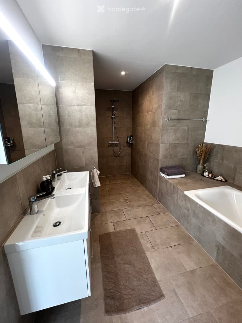 Modern bathroom with beige tiles, double sink, bathtub, shower, and decorative items including a vase with dried flowers and a towel on the edge of the bathtub.