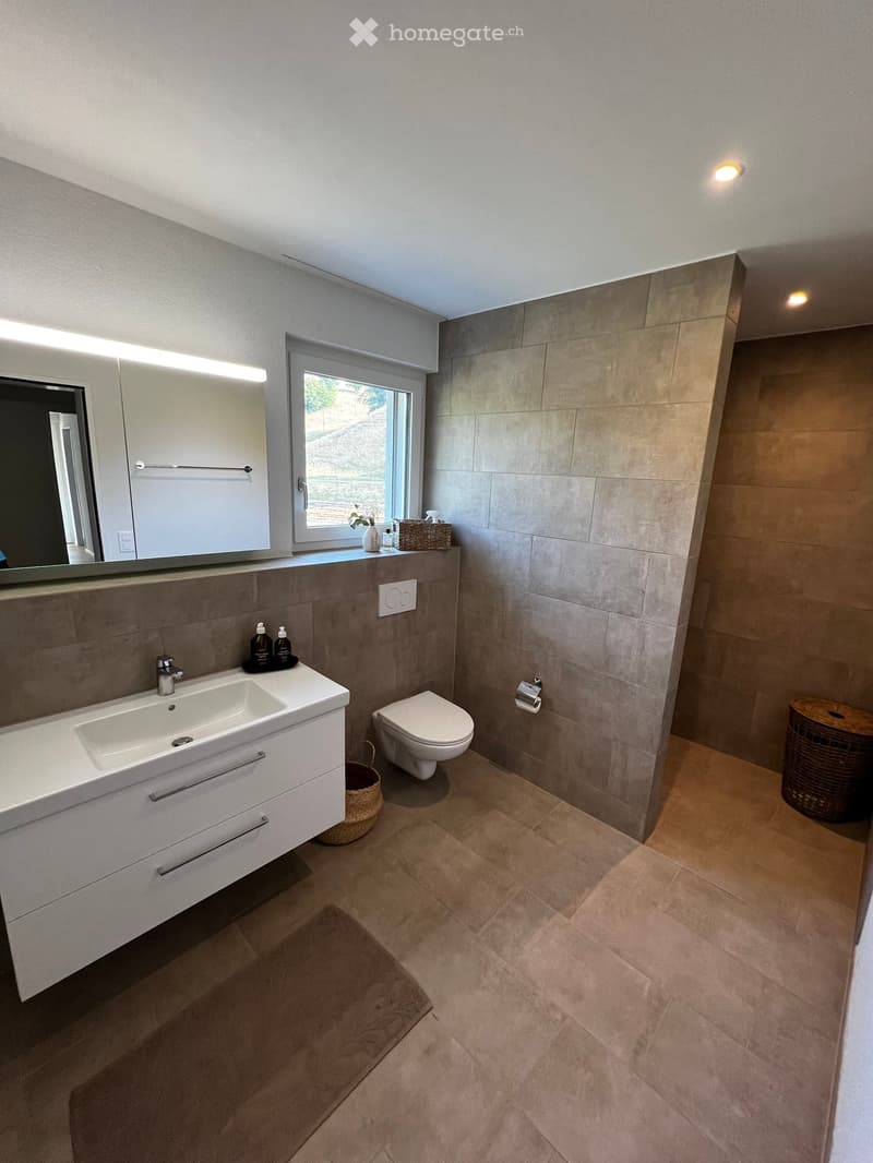 Modern bathroom with a white sink, toilet, large mirror, and wicker baskets. The walls are tiled in a neutral color, and the floor is made of beige tiles.