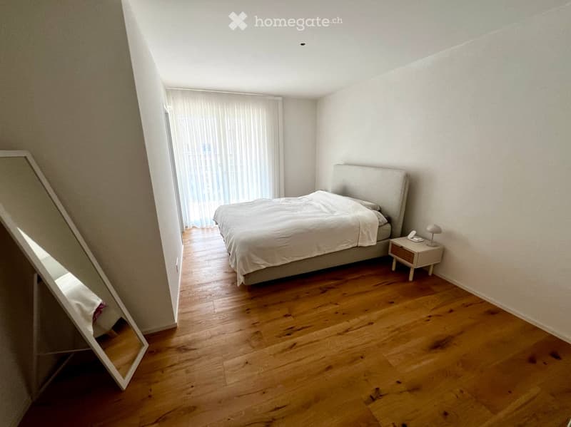 Modern bedroom with wooden flooring, large bed with white bedding, nightstand with lamp, and sheer curtains on the window.