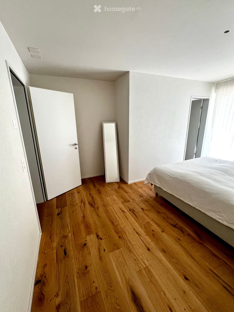 Bright bedroom with wooden flooring, a neatly made bed, a small mirror, and a closet door.