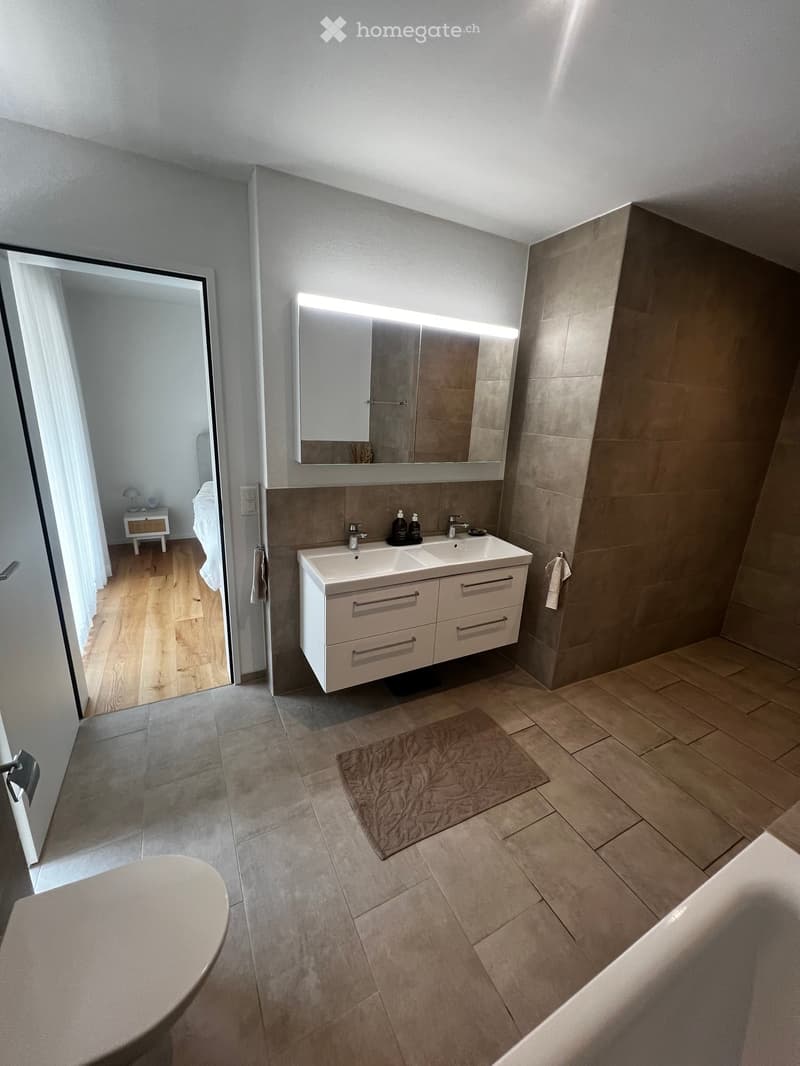 Modern bathroom with a double sink, large mirror, built-in cabinet, and a bathtub. The room features tiled floors and walls, a small rug, and an open doorway leading to a bedroom area with wooden flooring and a small nightstand.