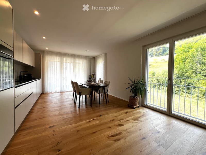 Open-plan kitchen and dining area with light wood flooring, modern kitchen cabinets, a dining table with chairs, and a large window leading to a balcony with a view of greenery.