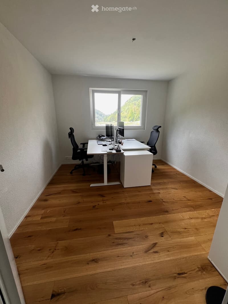 A modern, minimalist office space with a wooden floor, white walls, and a large window offering a scenic view. The room features a white desk with a laptop, monitor, and two ergonomic chairs.