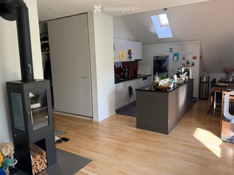 Modern kitchen with light wooden flooring, white cabinets, a black kitchen island, and a wood-burning stove. The space includes a sink, stove, fridge, and a dining area with a table and chairs.