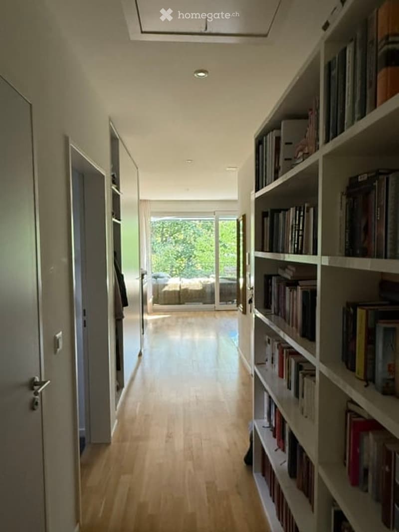 A modern, well-lit hallway with wooden flooring, built-in bookshelves, and a view into a living area through large glass doors.