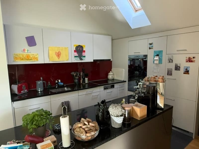 Modern kitchen with white cabinets, red backsplash, and stainless steel appliances. Features a central island with a bowl of bread, a vase, and a potted plant. The counter has a toaster, coffee maker, and other small appliances.