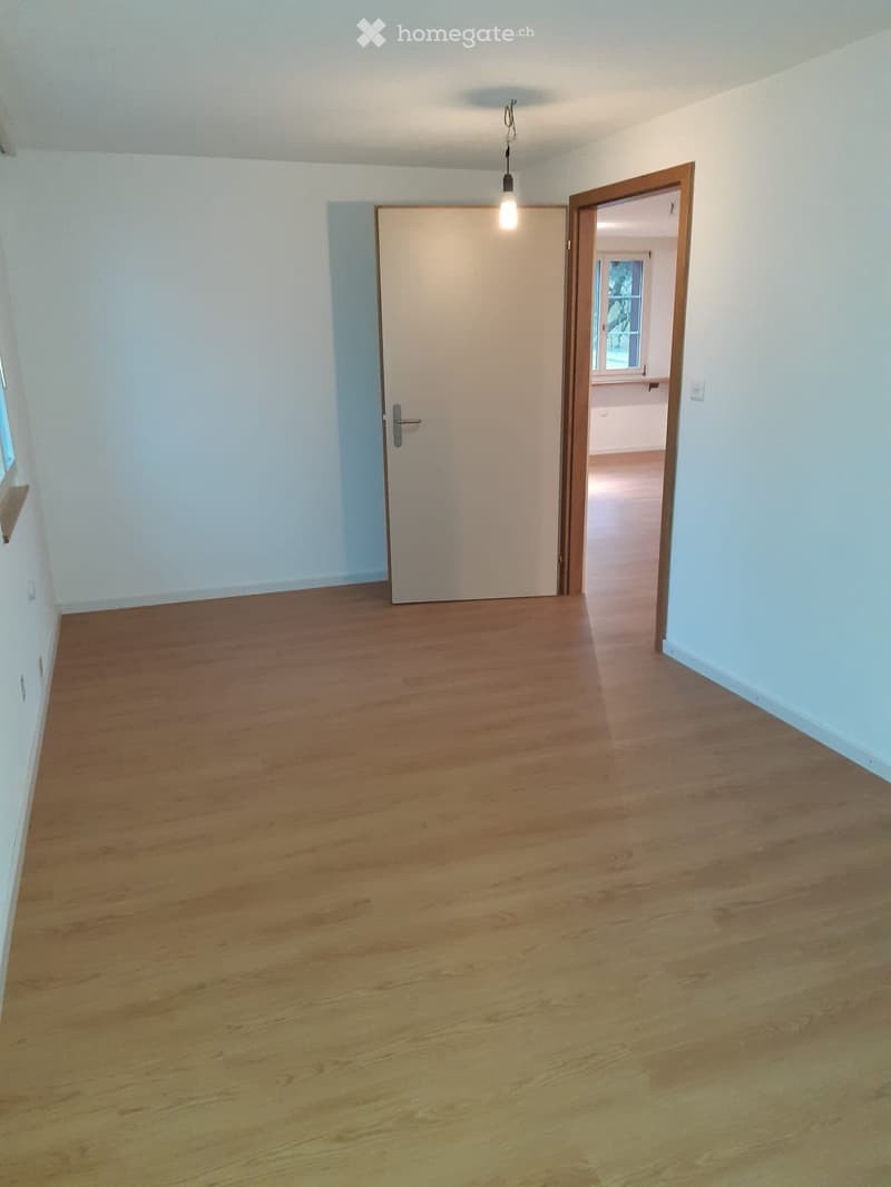 Empty hallway with wooden flooring, white walls, and two doors, one leading to another room with visible window and light fixture.