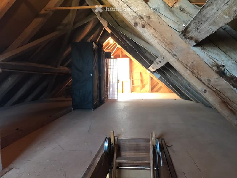 Empty attic space with wooden beams, exposed rafters, and a small opening to another room. The floor is unfinished, and there is a small staircase leading up.