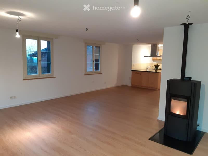 Empty room with wooden flooring, white walls, and a fireplace. Two windows with wooden frames are visible, and a small kitchen area is partially visible in the background.