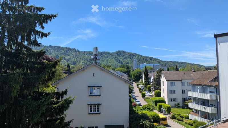 Residential street with multi-story houses, parked cars, and green surroundings with trees and hills in the background.