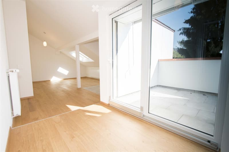Bright, empty room with wooden flooring, white walls, and large glass sliding doors leading to a balcony or outdoor area. Sunlight streams in, creating a clean and minimalist space.